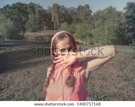 A girl with cheerful, playful, mischievous eyes covers her face with her hand. A cute young woman in pink clothes in the park, photo using filters and effects, in soft light pastel colors
