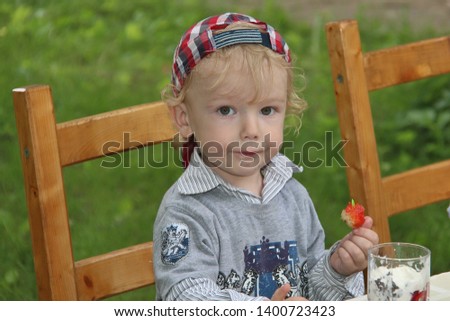 Blond 5-6 year old boy in cap eats strawberries with cream and ice cream. Cute little boy holding ice cream cone with strawberry jam in park. Chair stool and wooden table with food.