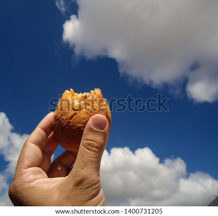 Holding a COXINHA DE FRANGO and having blue sky with cloud as background. The COXINHA DE FRANGO is a very popular food in Brazil. This image was produced in May 2019. Royalty-Free Stock Photo #1400731205