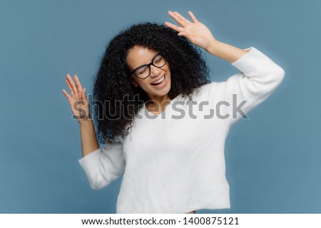 People and joy concept. Optimistic delighted Afro American woman raises hands, dances to loud music, being overjoyed, wears casual white jumper, isolated against blue background, has good mood