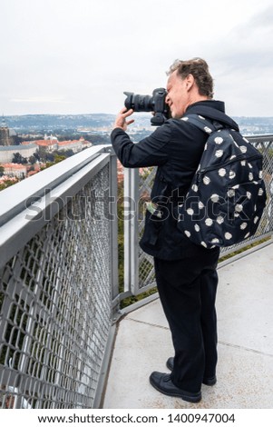 Outdoor profile view of a Caucasian male photographer in black suit and backpack at a tower with digital camera taking pictures of city view.