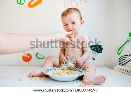 Dirty and smiling baby on a white table being fed by his mother's hand, while laughing while trying the blw method.