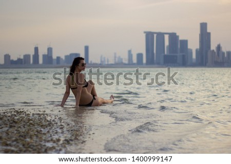Pretty girl is sitting in the shallow water with distant city in background during sunset