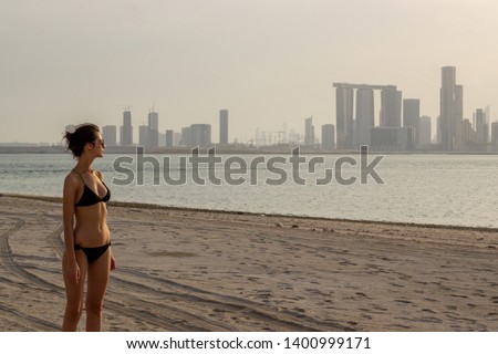 Pretty girl is standing at the beach with distant city in background during sunset