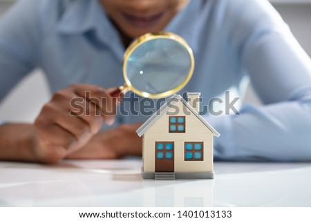 Close-up Of A Businesswoman's Hand Holding Magnifying Glass Over House Model Over Desk Royalty-Free Stock Photo #1401013133