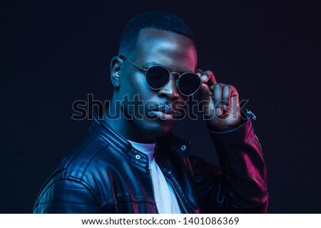 Young African male pictured against dark background in black jacket and trendy sunglasses