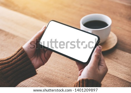 Mockup image of woman's hand holding black mobile phone with blank screen horizontally with coffee cup on wooden table
