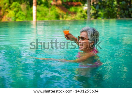 natural lifestyle portrait of attractive and happy middle aged Asian woman relaxed at tropical resort infinity simming pool with jungle background enjoying a drink relaxed in luxury holidays trip