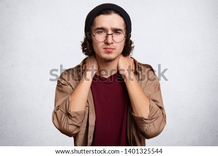 Indoor studio shot of handsome serious young guy posing isolated over white background in studio, touching his neck with both hands, doing massage, feeling pain aback, wearing casual clothes.
