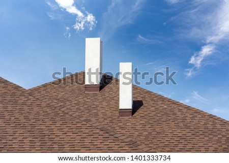 Close up photo of asphalt shingles or tiles installed on top of the new residential house under construction with two white chimneys, against blue sky background