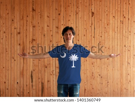 Man wearing South Carolina flag color shirt and standing with arms wide open on the wooden wall background, white palmetto tree on an indigo field and contains a white crescent. 