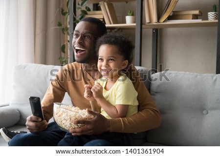 Happy African American father with toddler son, family watching tv, cartoons or football match, eating popcorn snack, sitting on couch together, little boy sitting on smiling dad knees at home