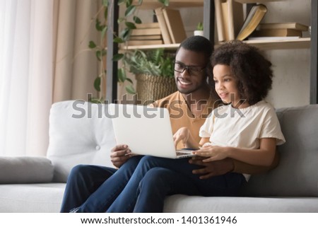 Smiling African American father in glasses with little daughter using laptop together, looking at screen, happy dad with preschool girl child watching cartoon or video, sitting on sofa at home