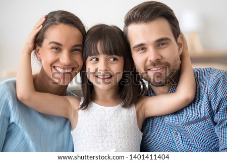 Family portrait of cute little daughter stand in between smiling young parents hugging them, happy mom and dad posing together with small girl child looking at camera laughing and cuddling