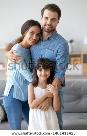 Portrait of happy young parents standing hug with cute preschooler daughter posing in living room, smiling loving family with little child girl embrace look at camera making picture at home together
