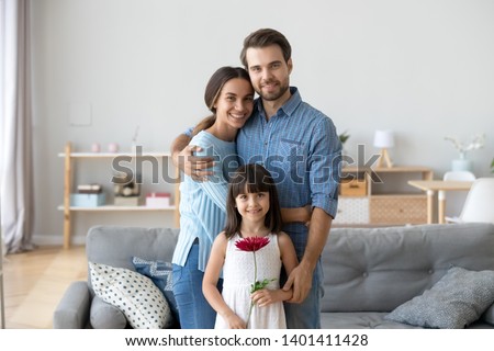 Portrait of happy young family with cute little child stand in cozy living room hugging look at camera, smiling parents embrace posing with preschooler daughter holding flower make picture at home