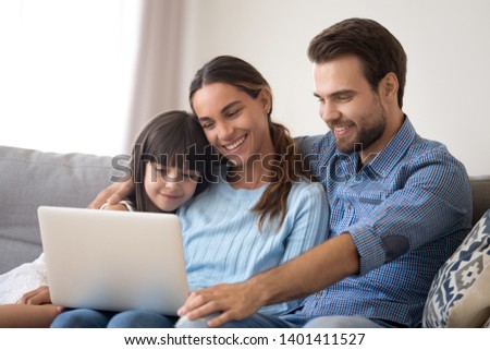 Happy young family with small kid sit relax cuddle on couch together watch movie on laptop, smiling parents rest on sofa enjoy spending weekend with preschooler daughter see cartoon on computer