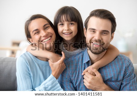 Family picture of smiling young mom and dad sit on couch posing with cute little daughter, small excited funny preschooler girl hug happy parents, relax on sofa look at camera making photo together