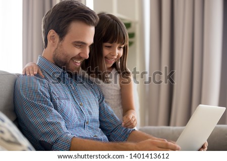 Cute little daughter hug young dad watch funny video on laptop laughing relaxing on couch together, sweet preschooler girl kid embrace father enjoy cartoon on computer spend time with parent at home