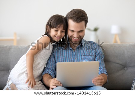 Happy young dad and preschooler daughter sit on couch watch cartoon on laptop together, smiling parent father enjoy time with little girl kid laugh on funny video on computer relaxing on sofa