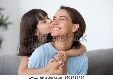 Cute small daughter kiss happy mom on cheek hugging her from behind, funny little caring preschooler kid piggyback embrace smiling mother showing love, child girl and mommy have sweet moment together