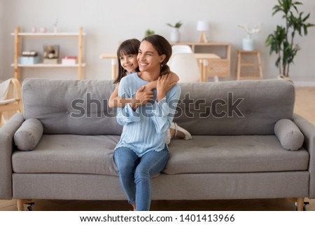 Cute little girl piggyback smiling young mother hugging from behind sit on cozy sofa in living room at home, happy mother and daughter embrace on sofa looking in distance dream of bright future Royalty-Free Stock Photo #1401413966