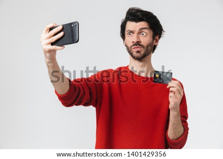 Handsome cheerful young bearded brunette man wearing sweater standing isolated over white background, taking a selfie, showing credit card