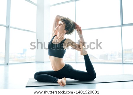 Young healthy flexible sportswoman in tracksuit holding her hands behind head while practicing stretching exercise in gym