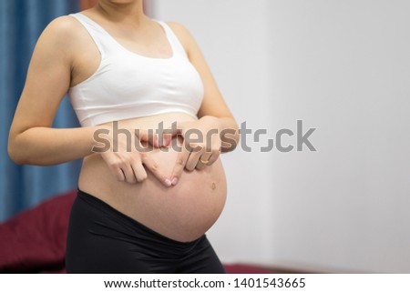 Pregnant woman standing in the bedroom wearing a white shirt, black pants, lifting hands, forming a heart shape.