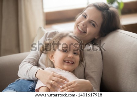 Portrait of happy young mom and cute preschooler daughter lying on couch hugging cuddling looking at camera, smiling mother or nanny embrace enjoy time with preschooler girl relaxing on sofa together