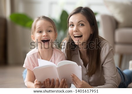 Happy young mother or nanny and funny little girl child lying on warm floor reading book together, smiling mom and preschooler daughter have fun flipping textbook look at camera laughing