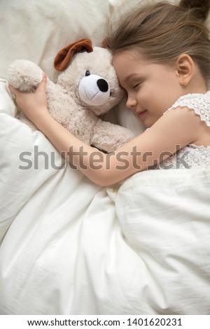 Cute little girl lying in cozy white bed hug plush teddy bear sleep peacefully seeing dreams, calm preschooler child fall asleep take nap with favorite toy rest on soft pillow on comfortable mattress