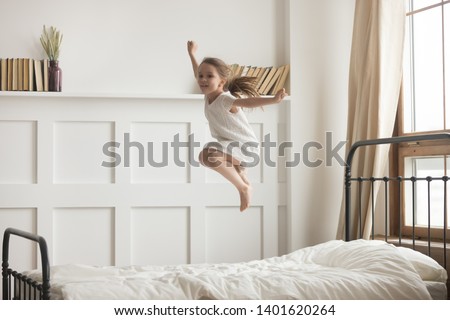Happy preschooler girl have fun jumping high on comfortable mattress on bed at home, excited little child entertain enjoy playing alone leap and hop in white bedroom engaged in funny activity Royalty-Free Stock Photo #1401620264