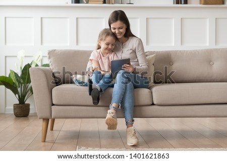 Happy young mom or nanny sit on couch relax with little daughter watch cartoon on tablet together, smiling mother and preschooler girl child have fun using playing on pad resting at home Royalty-Free Stock Photo #1401621863