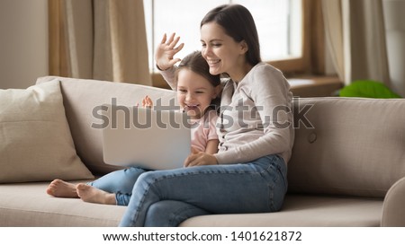 Smiling young mom and funny little daughter sit on couch hold laptop wave videochat talk on webcam, happy preschooler girl child and mother speak using computer have online conversation with dad