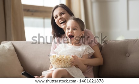 Happy young mom sit holding excited little daughter eating popcorn watch cartoon on TV together, smiling mommy or nanny have fun relaxing with small girl child enjoy movie or video with snack at home Royalty-Free Stock Photo #1401621878