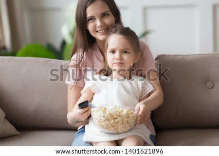 Funny little girl spend time with young mom watch TV eating popcorn relaxing at home together, cute small preschooler girl child hold remote search movie or cartoon rest with mommy on weekend