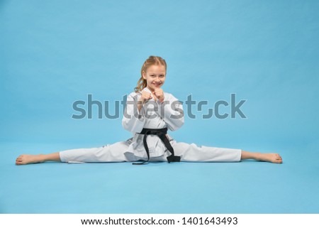 Karate girl in white kimono with black belt training, looking at camera and smiling. Positive, pretty child, making twine, sitting in combat position posing on blue background. 