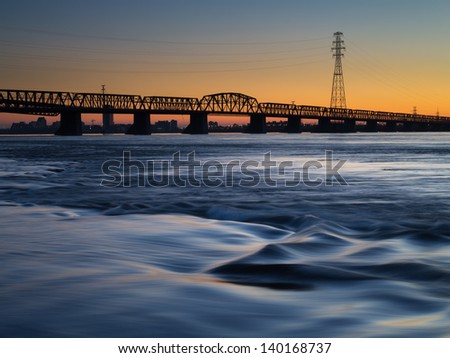 The sun sets over a bridge as a river flows in the foreground.
