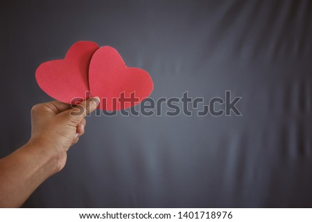 The woman's hand holds a pink paper heart with gray fabric background.