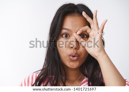 Close-up shot of intrigued and amazed enthusiastic young cute plump girl with tattoo showing ok or okay gesture over eye as peeking through hole at awesome thing, saying wow, folding lips curiously