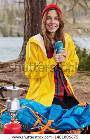 Outdoor shot of European woman with cheerful expression, stands on knees at ground in forest near river, holds flask of hot drink, wears red hat and yellow raincoat, coffee maker on camping stove