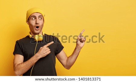 Horizontal shot of hipster guy points away with both fore fingers, wears stylish hat and casual black t shirt, has headphones on neck for listening audio tracks, isoated over yellow background