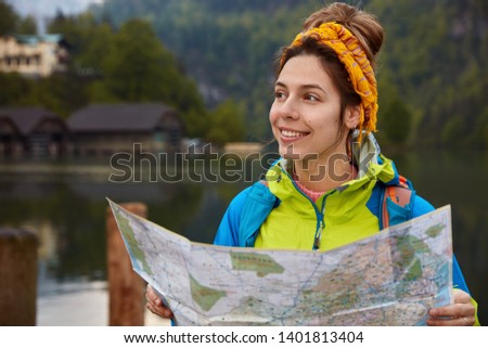 Positive young woman with joyful expression, holds location map, stands against beautiful nature with river, small houses and forest, enjoys leisure, dressed in anorak and headband, has smile on face