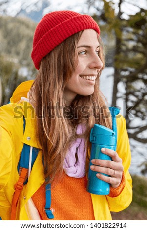 Vertical shot of smiling female traveler wears red hat, yellow coat, stretches hand, makes selfie with unrecognizable device, holds blue thermos, has pleasant smile, poses over pine forest outdoor