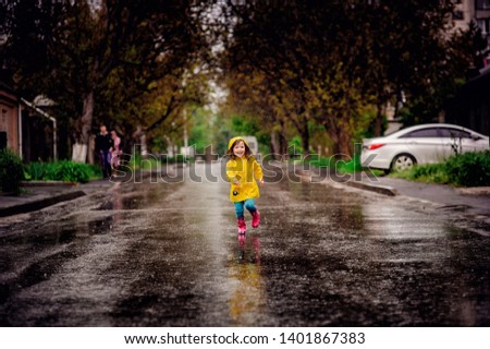 Little toddler girl jumping over puddle. Girl under the rain wearing yellow raincoat and pink rubber boots. Walk on street child.