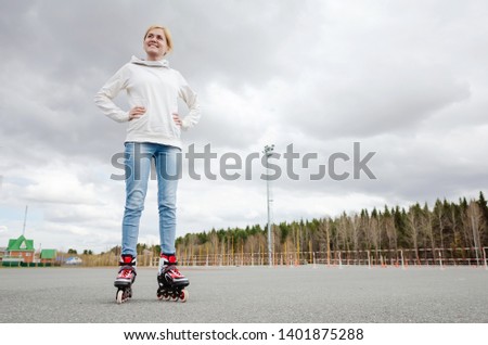 A young blond girl in a white sweatshirt and blue jeans, casual, roller skating in the stadium. The concept of summer activity, healthy lifestyle, summer fun. The girl smiles.