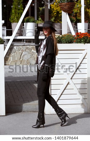 Fashionable girl dressed in black trousers, a stylish gray shirt and sweater and in a black hat with wide brim walks in the park on the sunny day
