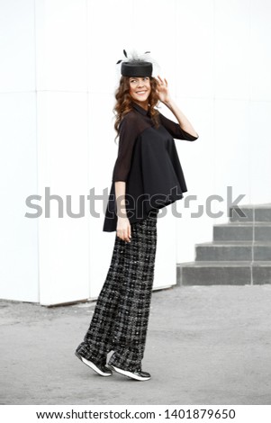 Brunette girl dressed in a stylish black shirt, flared pants and fashionable little hat poses in the street on a sunny day in the city