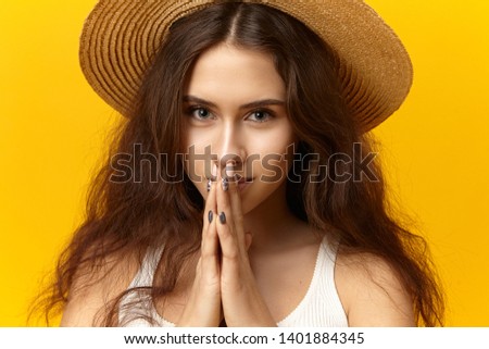 Human facial expressions and body language. Picture of beautiful gorgeous young woman with loose voluminous hair posing at yellow wll in straw hat, holding hands in prayer, having hopeful look